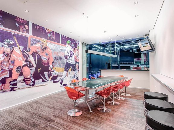 Die komplette linke Wand ist mit einem Poster von Spielern bei einem Eishockey-Spiel verziert. In der Mitte steht ein gläserner, länglicher Tisch mit Stühlen dran. Dahinter ist der Zugang zum Innenraum. 