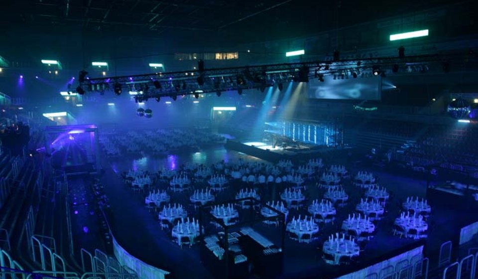Blick aus dem Oberrang in den Innenraum. Auf der abgedeckten Fläche stehen mehrere eingedeckte Tische um eine Bühne herum. Auf die Bühne scheinen Scheinwerfer, die Halle ist blau beleuchtet. 
