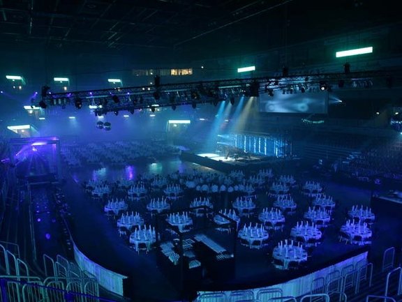 Blick aus dem Oberrang in den Innenraum. Auf der abgedeckten Fläche stehen mehrere eingedeckte Tische um eine Bühne herum. Auf die Bühne scheinen Scheinwerfer, die Halle ist blau beleuchtet. 