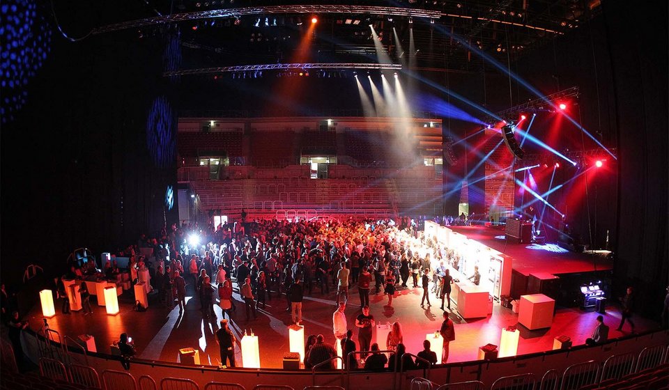 Auf der abgedeckten Fläche ist eine Bühne aufgebaut, vor der eine längliche Theke steht. Von der Bühne strahlen rote und blaue Lichtkegel durch den Raum. Im abgedunkelten Raum stehen viele Menschen auf der Fläche unmittelbar vor der Theke. 